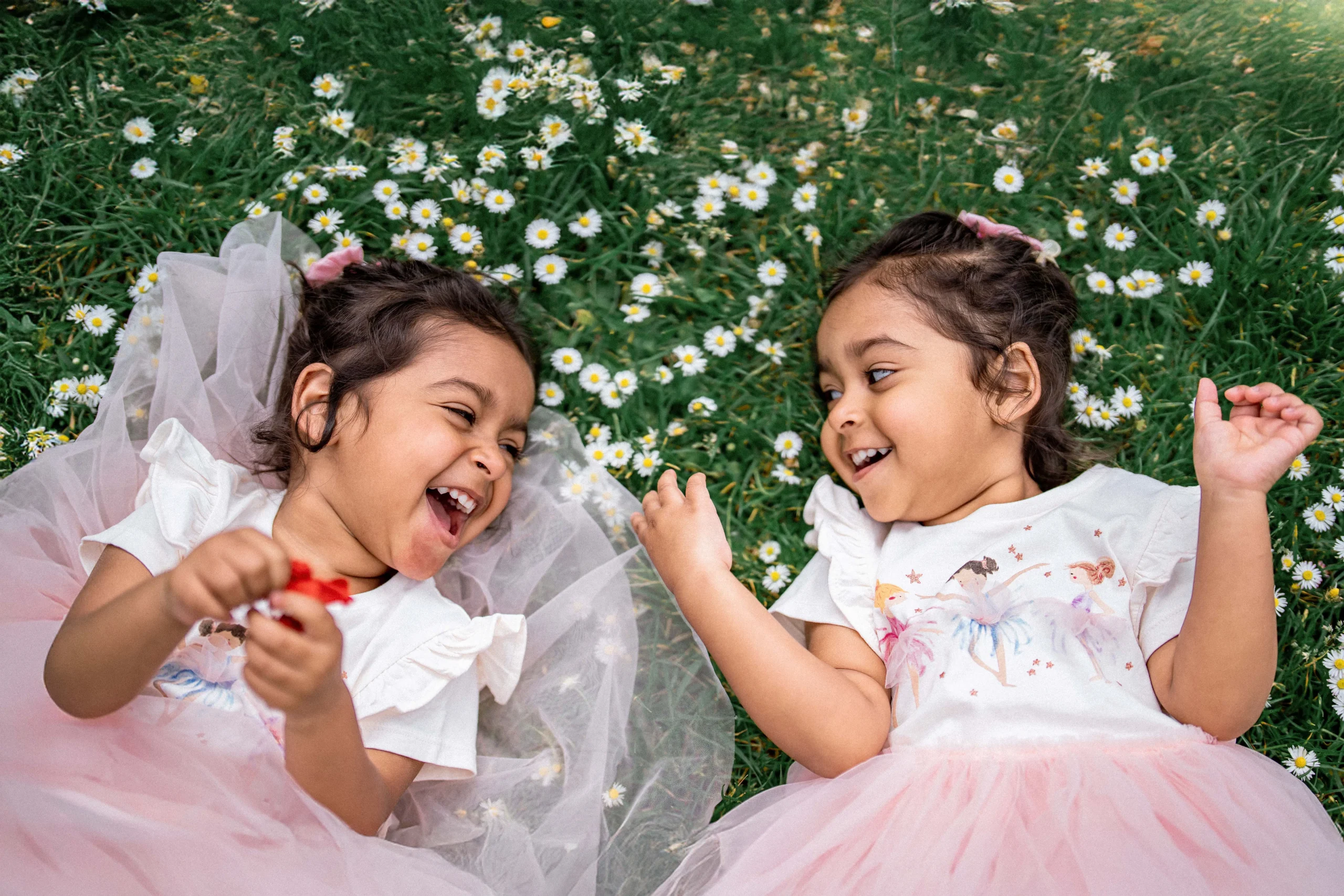 Two happy young girls laughing while lying on green grass with white daisies in a London garden, professional family photography by London Snap.