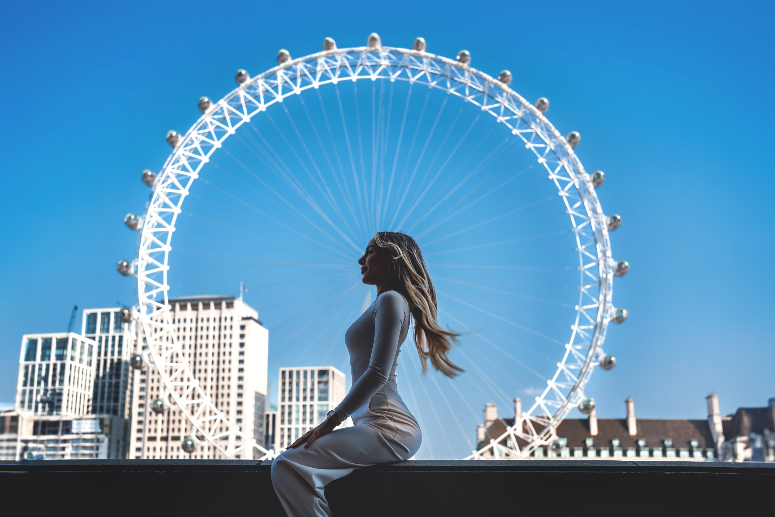 A solo female traveler sitting elegantly in front of the iconic London Eye against a clear blue sky, professional lifestyle photography by London Snap.