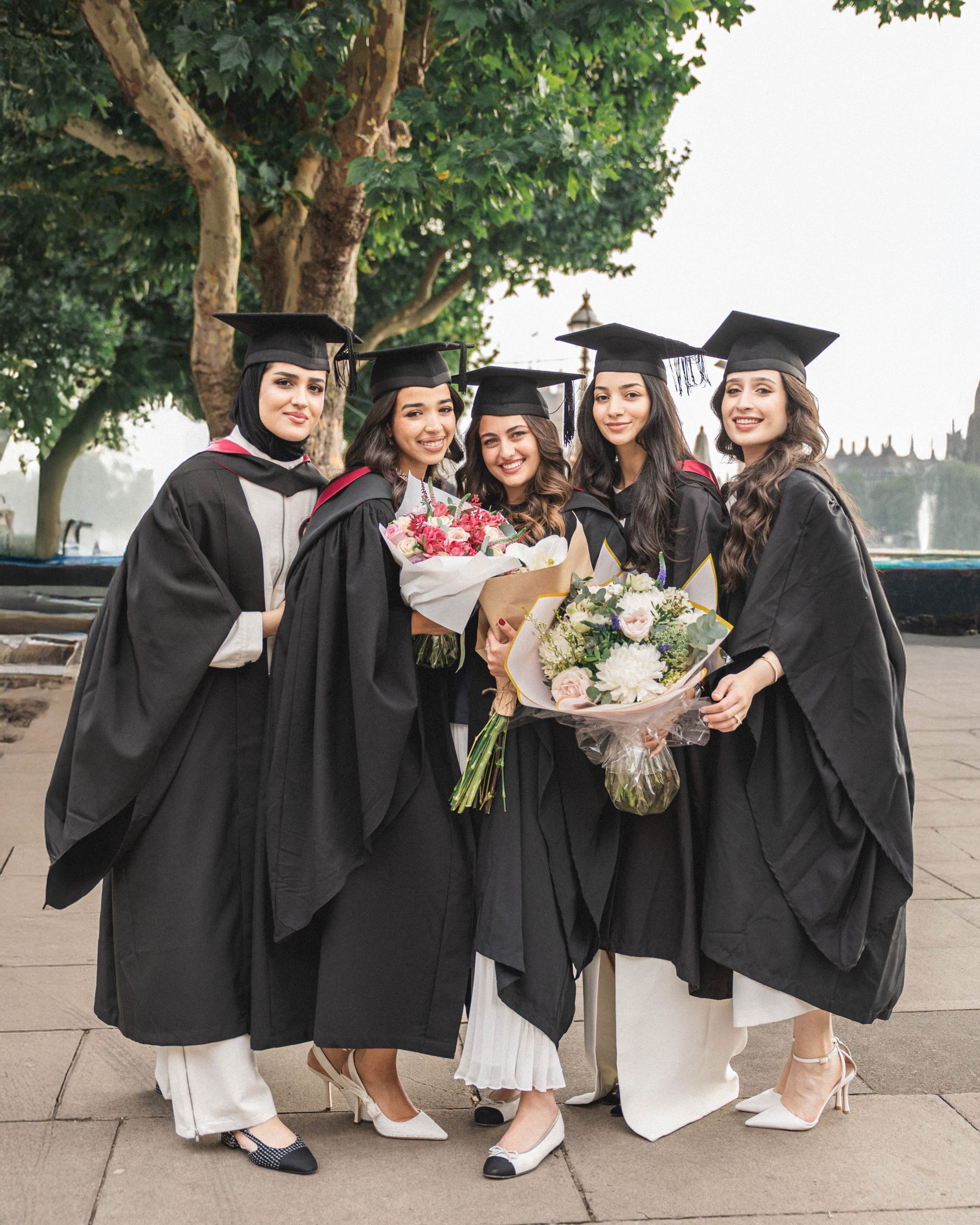 Five diverse female graduates in caps and gowns holding bouquets of flowers and smiling at a London university campus, captured by London Snap photography agency.
