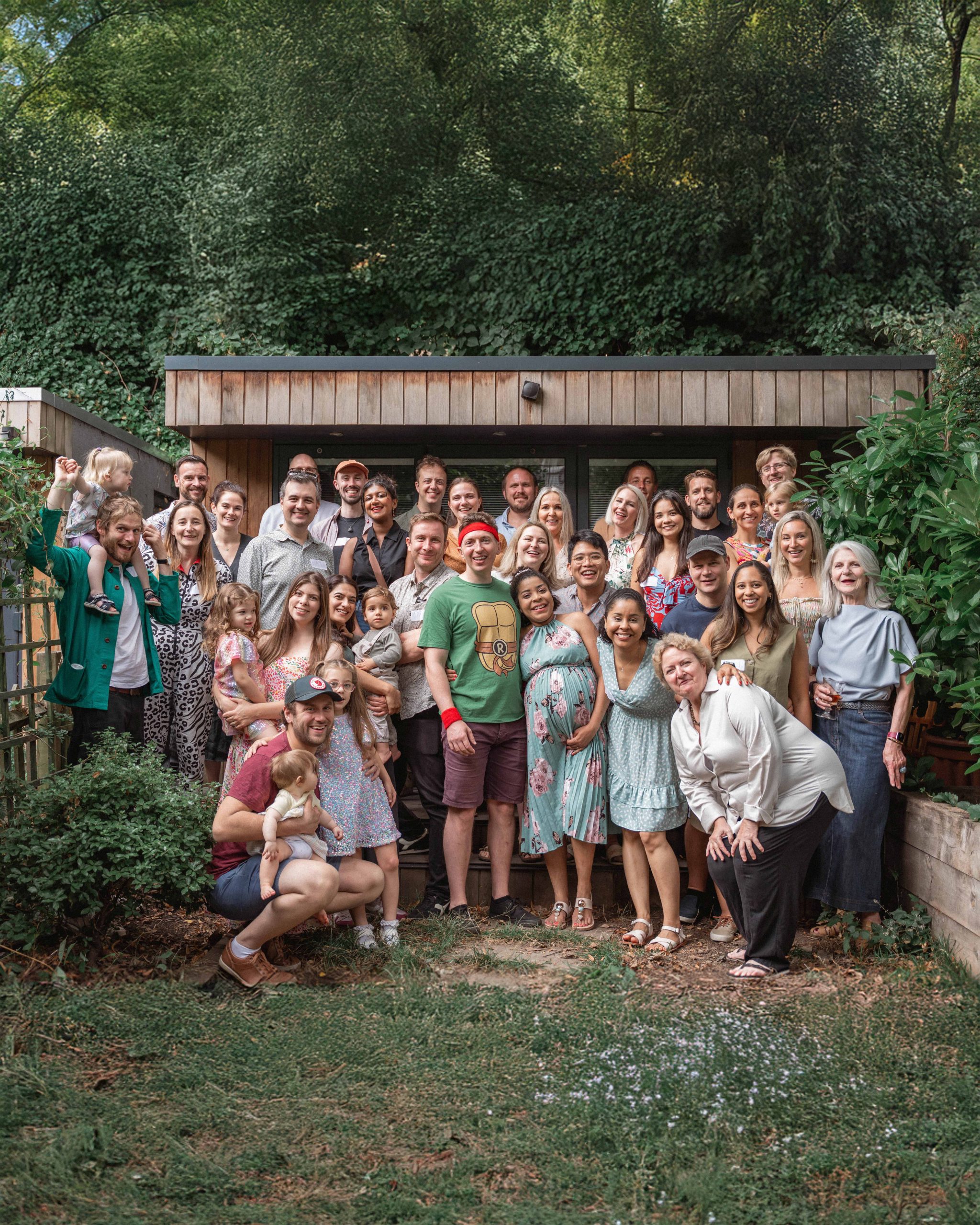 A beautiful maternity portrait of a woman in a floral dress smiling alongside a large group of family and friends in a lush green London garden, captured by London Snap.