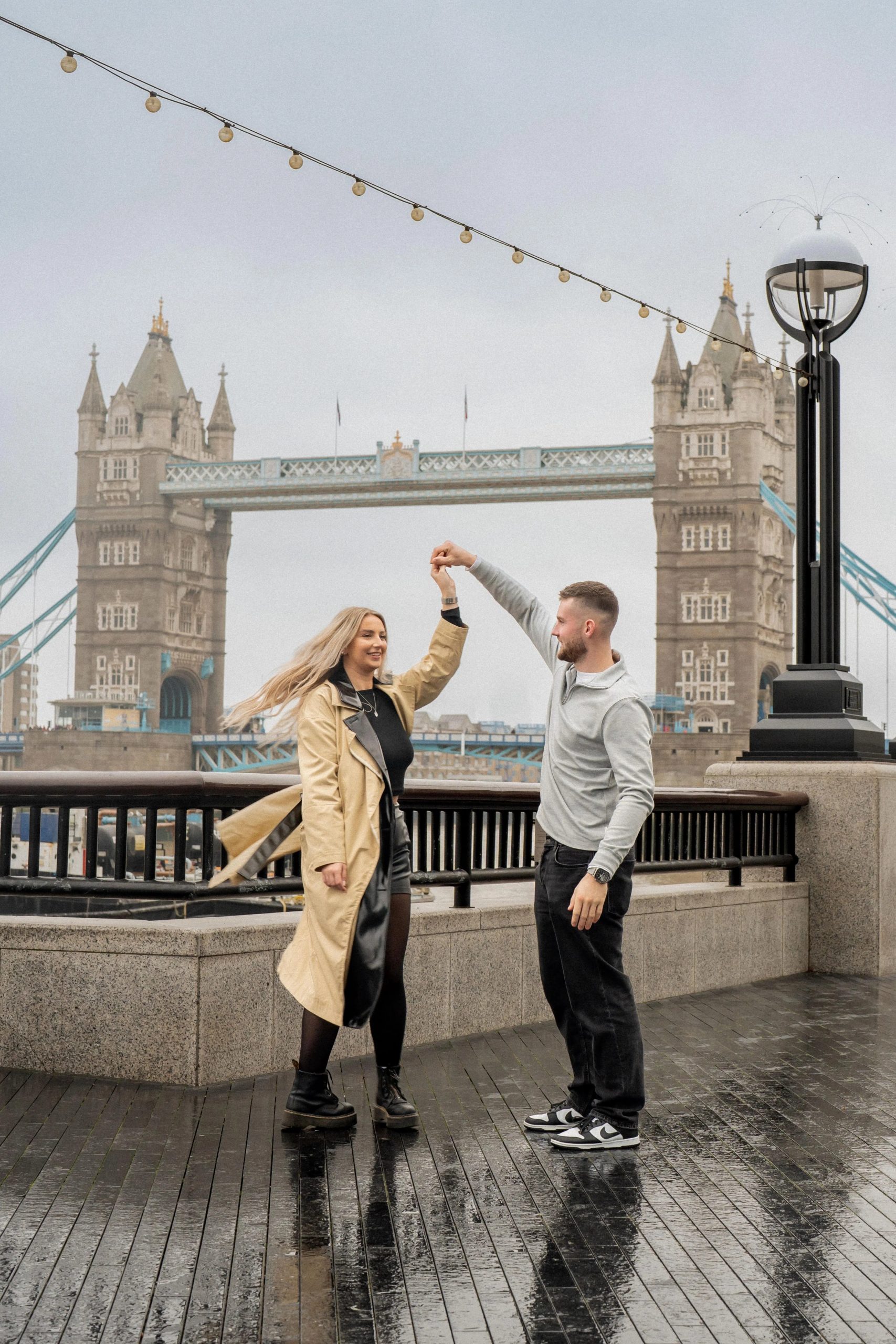 A romantic couple dancing and twirling in front of the iconic Tower Bridge on a rainy London day, professional photography by London Snap.
